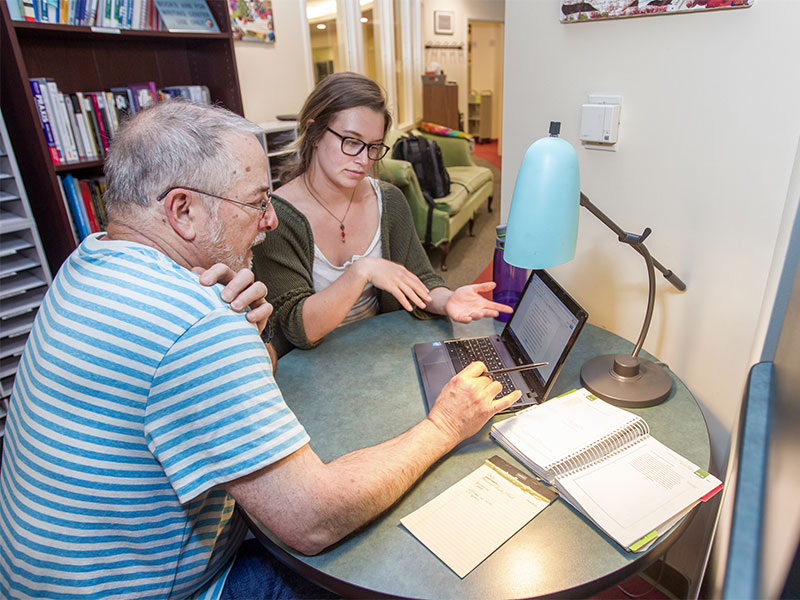 Two people sit at a round table discussing work on a laptop in a study space.