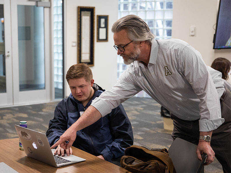 An advisor leans over a table to point at a student’s laptop screen during a one‑on‑one meeting.