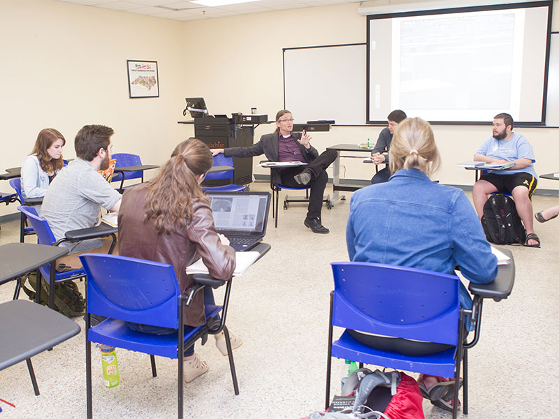 Students sit in a classroom arranged in a circle, listening and taking notes during discussion.”