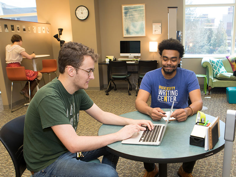 Two people sit at a small table in a writing center, working together on a laptop.