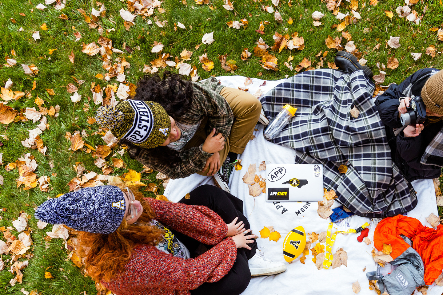 Three people sit on a blanket amid autumn leaves, chatting and taking photos during an outdoor gathering.