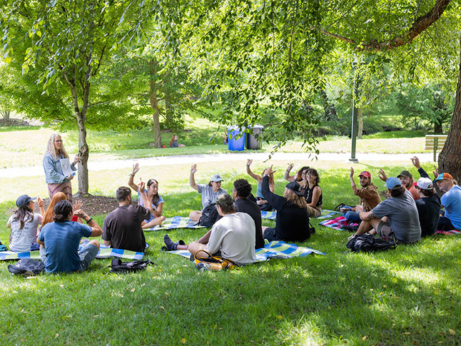 A group of students sits in a circle on the grass during an outdoor class, raising their hands to participate.